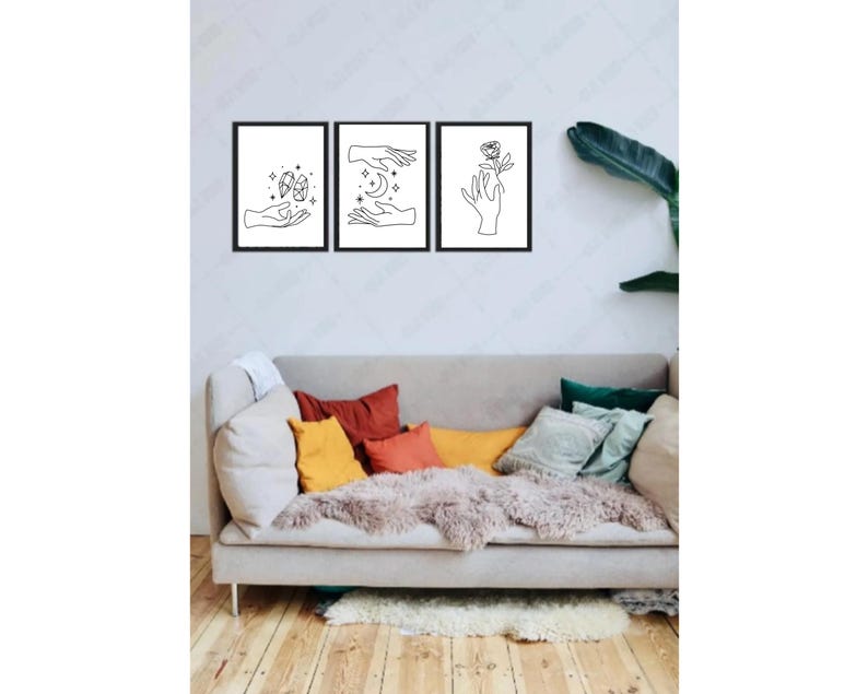 May include: Three black framed prints with line drawings of hands holding a crescent moon, a flower, and a star. The prints are hanging on a white wall above a light gray couch with colorful throw pillows.