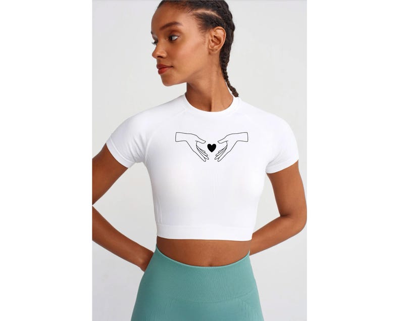 May include: White crop top with short sleeves featuring a black line drawing of two hands holding a heart in the center.