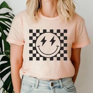 Może przedstawiać: R&oacute;żowy t-shirt z czarno-białym wzorem w kratkę i czarną buźką z błyskawicami zamiast oczu.