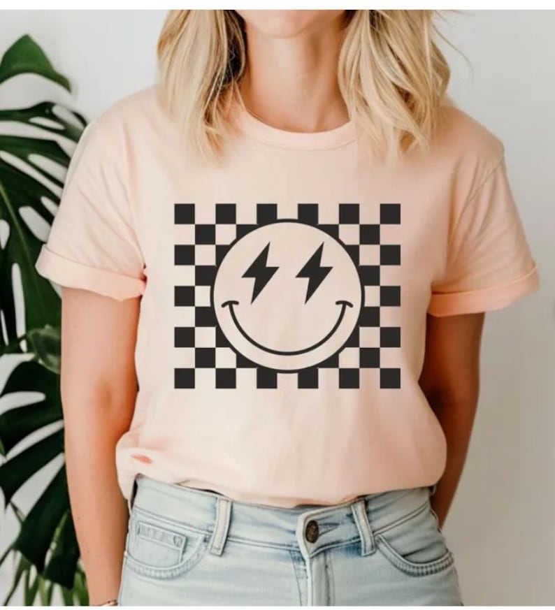 Może przedstawiać: R&oacute;żowy t-shirt z czarno-białym wzorem w kratkę i czarną buźką z błyskawicami zamiast oczu.