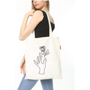 May include: A white tote bag with a black line drawing of a hand holding a rose. The bag has a red label on the side.
