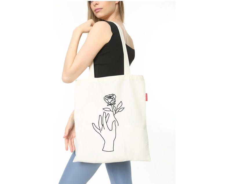 May include: A white tote bag with a black line drawing of a hand holding a rose. The bag has a red label on the side.