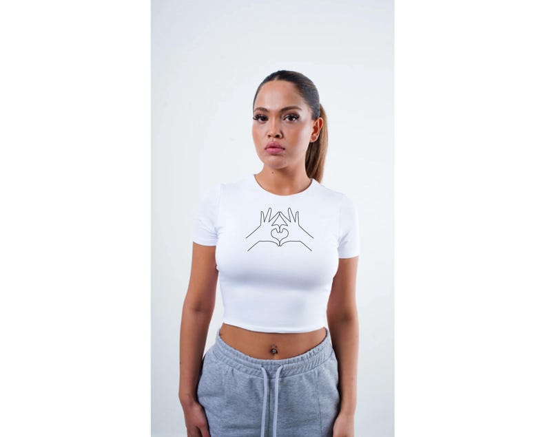 May include: White crop top with a black line drawing of two hands making a heart shape.