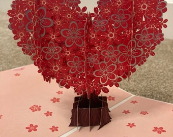 May include: A three-dimensional pop-up greeting card featuring a heart-shaped tree structure made of layered red paper flowers with silver outlines. The base of the tree is dark brown with a wood grain pattern, and the flat card surface is light pink.