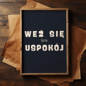 May include: A framed print featuring the Polish phrase "WEŹ SIĘ kurwa USPOKÓJ" in white, bold lettering on a dark blue backdrop. The frame is light brown, and the print is set against brown paper and a wooden surface.