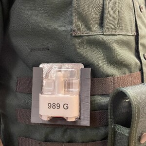 May include: A black and gray pouch with a clear plastic window containing a small device with the text "989 G". The pouch is attached to a green tactical vest with black straps.