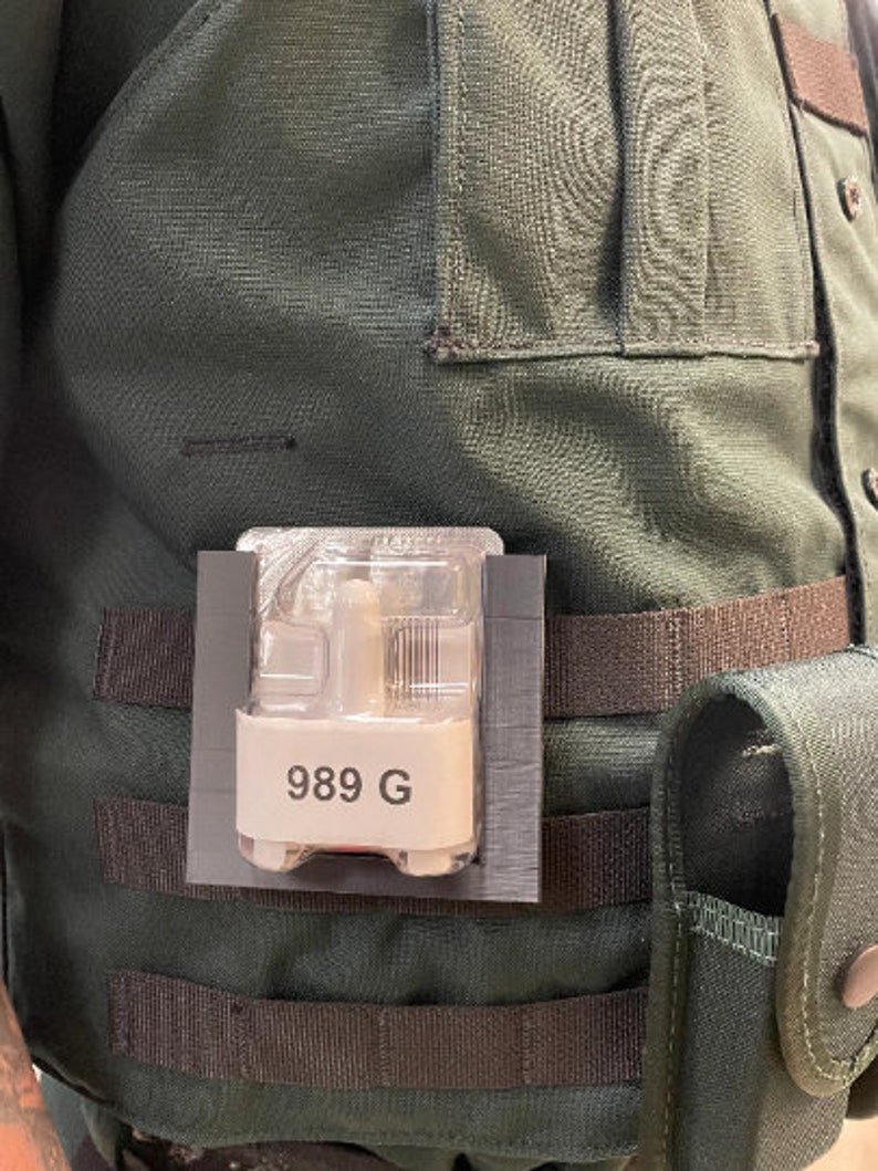 May include: A black and gray pouch with a clear plastic window containing a small device with the text "989 G". The pouch is attached to a green tactical vest with black straps.