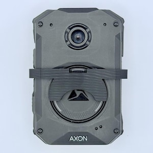 May include: A gray rectangular device with a black strap, a central lens, and the word "AXON" printed at the bottom. The device has a textured surface and appears to be a body camera or similar recording device.