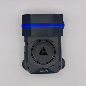 May include: A black device with a blue stripe and the word "AXON" printed on the bottom. The device has a circular center with a triangle logo. The top portion is a black, geometric shape.
