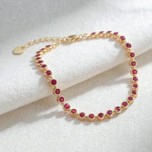 May include: A gold-colored bracelet featuring a series of round, red gemstones. The bracelet is set on a white surface, and the clasp is visible. The bracelet is a delicate piece of jewelry.