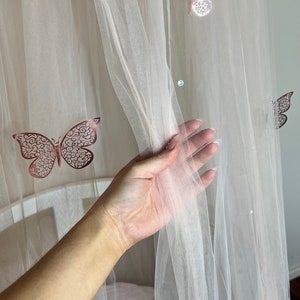 Könnte beinhalten: Ein weißer, durchsichtiger Baldachin mit einer Hand, die den Stoff hält. Der Baldachin hat ein rosafarbenes Schmetterlingsdesign.