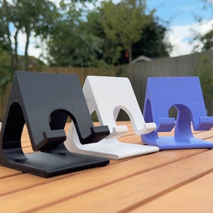 May include: Three phone stands made of plastic in black, white, and purple. The stands are angled and have a curved lip to hold a phone.