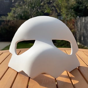 May include: A plain white mask with a curved top and eye openings. The mask features a beak-like shape and is made of a smooth, matte material. It is designed to be worn on the face, suitable for costumes or artistic projects.