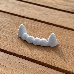 May include: A set of white plastic vampire teeth. The teeth are arranged in a curved row, with two pointed fangs on either end and three smaller teeth in the middle.