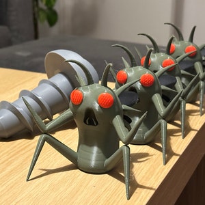Puede incluir: Un conjunto de cinco figuritas de insectos verdes impresas en 3D con ojos rojos. Los insectos están dispuestos en línea, con el insecto más grande en la parte posterior y el más pequeño en la parte delantera. Los insectos están todos mirando en la misma dirección. Los insectos están sentados sobre una superficie de madera.