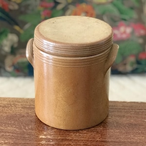 May include: A small, round, tan-colored stoneware jar with a lid. The jar has two small handles and a ridged design around the top and bottom. The lid is flat and slightly lighter in color than the body of the jar. The jar is sitting on a wooden surface.