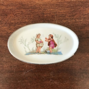 May include: Oval porcelain dish with a silver rim featuring a vintage illustration of two girls in a garden setting. One girl is wearing a green dress and the other is wearing a pink dress.