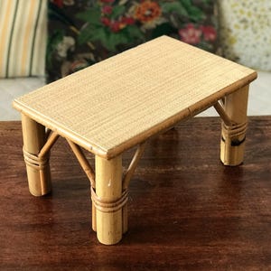 Può includere: Un piccolo tavolo rettangolare in bambù con piano intrecciato. Il tavolo ha quattro gambe, ciascuna realizzata con pali di bambù legati con materiale intrecciato. Il tavolo è di colore marrone chiaro naturale.