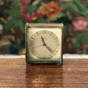 Op de afbeelding: Een vintage zilverkleurige wekker met een witte wijzerplaat en zwarte wijzers. De klok heeft een vierkante vorm en een glazen voorzijde. De wijzerplaat van de klok heeft zwarte Romeinse cijfers als uurmarkeringen en zwarte minutenmarkeringen.