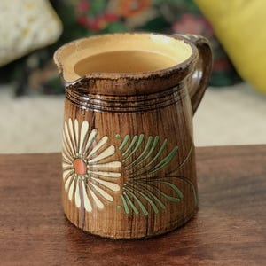 May include: A brown ceramic pitcher with a floral design. The pitcher has a handle and a spout. The floral design features a white daisy with an orange center and green leaves. The interior is a light yellow color.