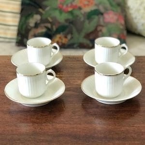 Op de afbeelding: Vier witte espresso kopjes en schotels. Elk kopje heeft een gouden rand en een verticaal geribbeld ontwerp. De schotels zijn wit met een gouden rand en een subtiel, stralend patroon. De set staat op een donker houten oppervlak.