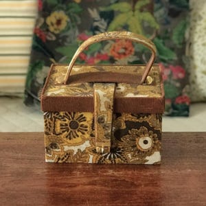 Könnte beinhalten: Eine Vintage-Handtasche mit floralem Muster in Braun-, Gelb- und Cremetönen. Sie hat einen Griff und einen Schnallenverschluss. Die Seiten sind mit einem dunkleren braunen Stoff verkleidet. Die Tasche steht auf einer Holzoberfläche.