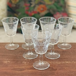 May include: A collection of seven clear glass cordial glasses, each featuring an etched floral design. The glasses have a stemmed base and a flared bowl. They are displayed on a wooden surface, with a patterned cushion in the background.