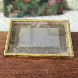 Może przedstawiać: Mała, prostokątna ramka na zdjęcia z wyblakłą, żółtą, pomalowaną drewnianą ramą. Ramka zawiera jasny obraz, prawdopodobnie zdjęcie lub grafikę. Ramka leży na ciemnej drewnianej powierzchni.