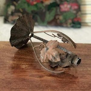 Può includere: Un portalampada vintage in metallo marrone arrugginito con dettagli di foglie e viti. L'apparecchio ha un grande elemento a forma di ventaglio e un portalampada per una lampadina. Il design ricorda l'arredamento antico.