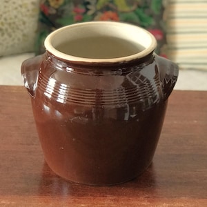 May include: A brown ceramic pot with a glossy finish. The pot has a wide opening with a cream-colored rim and two small handles. Horizontal ridges encircle the upper portion of the pot. The pot is sitting on a wooden surface.