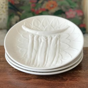May include: A stack of three white ceramic plates with an embossed asparagus design. Each plate has a central trough and a floral pattern around the edge. The plates are stacked on a wooden surface.