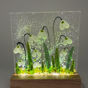 May include: A glass panel depicting snowdrop flowers with white petals and green stems. The panel is set on a wooden base with an integrated light. The flowers are set against a speckled background, creating a soft, illuminated display.