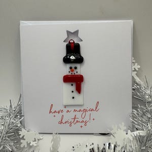 May include: A square white greeting card featuring a fused glass snowman ornament with a black top hat, red scarf, and orange nose. The card has the red printed text "have a magical christmas!" with small red plus signs and a star cutout above the hat.