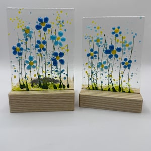 May include: Two rectangular glass art pieces featuring blue and yellow floral designs. The glass panels are mounted on wooden bases. The flowers have blue petals and yellow centres, with green stems and leaves. The background is white.