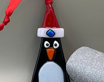 May include: A glass penguin Christmas ornament with a red Santa hat, white trim, and a blue gem. The penguin has a black body, white belly, and orange beak and feet. A red ribbon is attached for hanging.