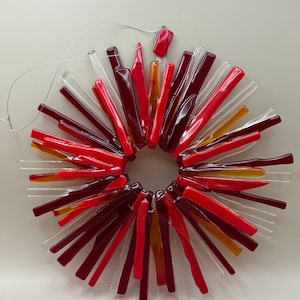 May include: A circular glass ornament with a sunburst design. It features glass rods in red, burgundy, clear, and yellow. A thin wire is attached for hanging. The ornament is approximately 15 cm in diameter.