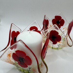 May include: Two clear glass candle holders with wavy, petal-like edges, each decorated with vibrant red poppy flower designs and green stems. A solid white pillar candle sits inside the front holder, which features a red rim and orange speckles.