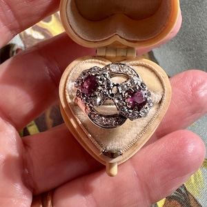 May include: A silver-toned ring featuring two prominent round-cut purple stones surrounded by small clear stones, presented inside a small, cream-colored, heart-shaped hinged box with a velvet-lined interior.