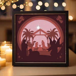 Könnte beinhalten: Gerahmtes Papierkunstwerk mit einer Darstellung der Geburt Christi. Die Kunst zeigt Silhouetten von Figuren, Palmen und einem Stern in Burgunder- und Rosatönen. Der schwarze Rahmen umgibt die Szene, die von einem warmen Licht beleuchtet wird.