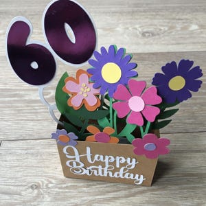 May include: A handmade birthday card featuring a pop-up floral arrangement with purple, pink, and orange flowers. The card includes the number 60 in metallic purple and the words "Happy Birthday".