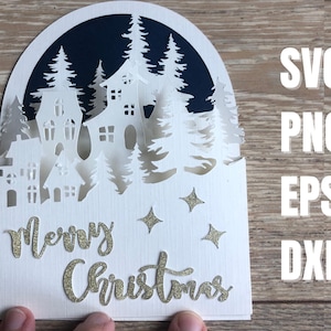 May include: A layered paper Christmas card featuring a winter village scene with houses and trees, cut from white paper. The words "Merry Christmas" are in gold glitter script. The card also includes the file types SVG, PNG, EPS, and DXF.