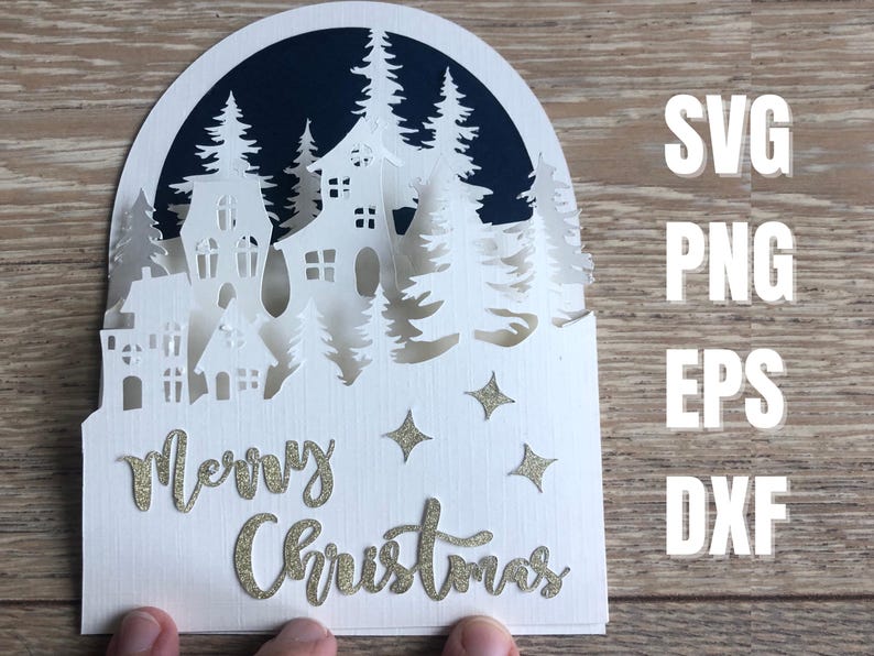 May include: A layered paper Christmas card featuring a winter village scene with houses and trees, cut from white paper. The words "Merry Christmas" are in gold glitter script. The card also includes the file types SVG, PNG, EPS, and DXF.