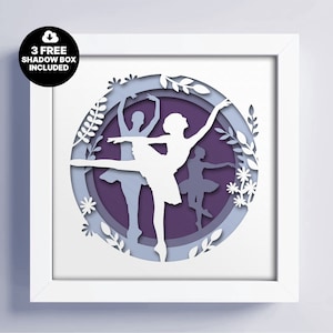 Può includere: Una cornice bianca con un design di carta tagliata in viola, blu e bianco di tre ballerine in un cerchio con accenti floreali. Il testo "3 FREE SHADOW BOX INCLUDED" è in un cerchio verde nell'angolo in alto a sinistra dell'immagine.