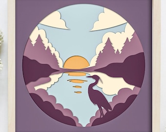 Puede incluir: Una obra de arte recortada en papel que representa una garza de pie junto a un lago con una puesta de sol en el fondo. La escena está enmarcada por un círculo y se coloca sobre un fondo morado.