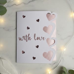 Pu&ograve; includere: Un biglietto bianco con la scritta "with love" in glitter oro rosa. Il biglietto presenta ritagli a forma di cuore e piccoli accenti a cuore. Luci fatate e verde circondano il biglietto.