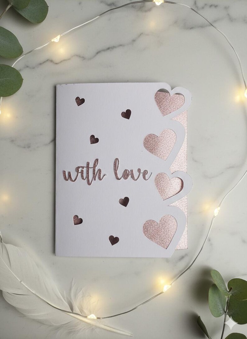 Pu&ograve; includere: Un biglietto bianco con la scritta "with love" in glitter oro rosa. Il biglietto presenta ritagli a forma di cuore e piccoli accenti a cuore. Luci fatate e verde circondano il biglietto.