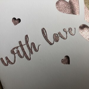 Pu&ograve; includere: Un biglietto bianco con la scritta "with love" in oro rosa glitterato. Il biglietto presenta forme di cuore ritagliate e piccoli ritagli a forma di cuore, creando un design romantico ed elegante.