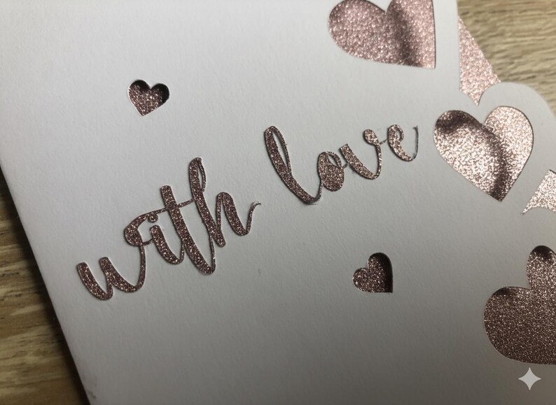 Pu&ograve; includere: Un biglietto bianco con la scritta "with love" in oro rosa glitterato. Il biglietto presenta forme di cuore ritagliate e piccoli ritagli a forma di cuore, creando un design romantico ed elegante.