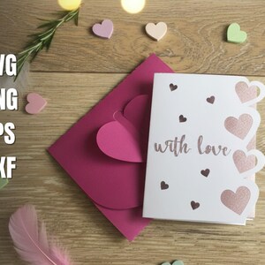 Pu&ograve; includere: Un biglietto di auguri bianco con la scritta "with love" in corsivo, con ritagli a forma di cuore e piccoli accenti a cuore. Sono presenti anche una busta rosa e cuori decorativi, su uno sfondo di legno.