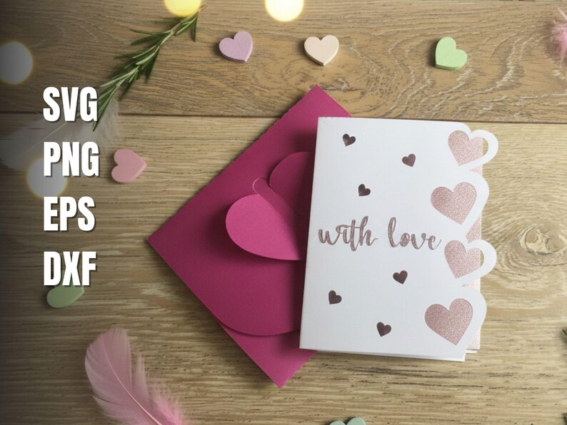 Pu&ograve; includere: Un biglietto di auguri bianco con la scritta "with love" in corsivo, con ritagli a forma di cuore e piccoli accenti a cuore. Sono presenti anche una busta rosa e cuori decorativi, su uno sfondo di legno.
